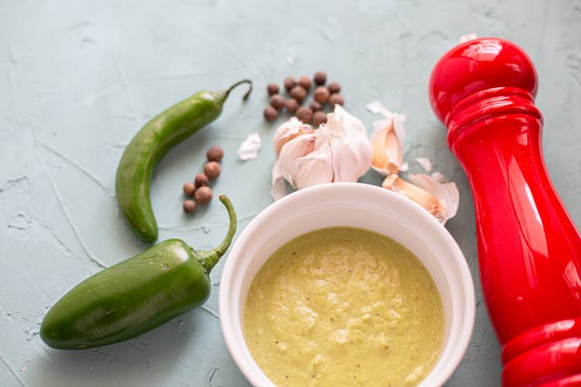 photo of a bowl of soup, green chili peppers, beans, a head of garlic, and a red pepper mill